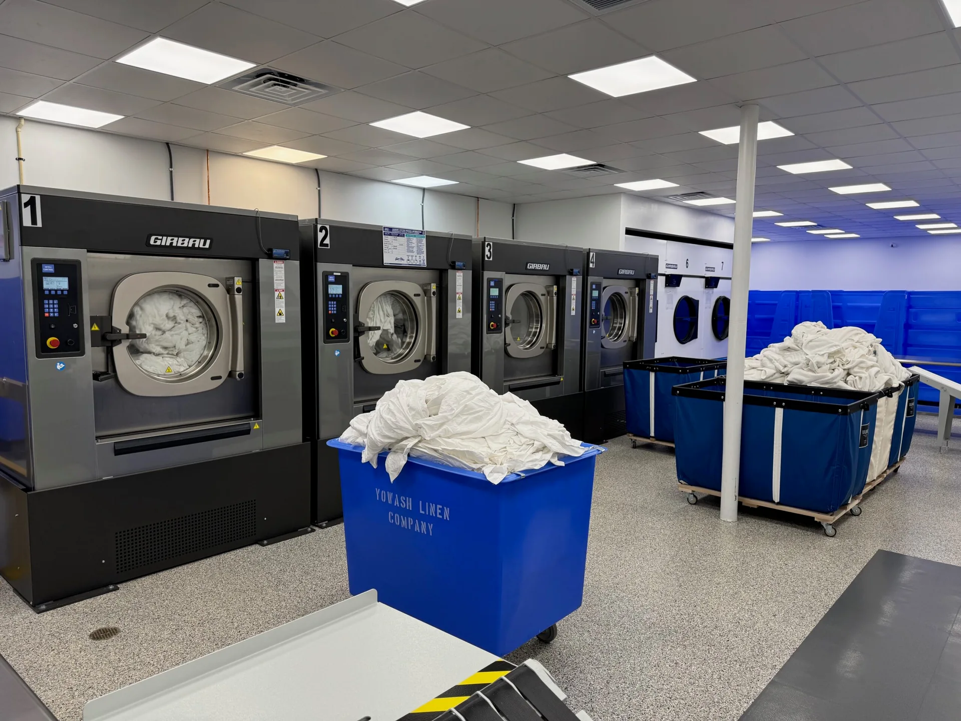 Commercial washing machines and blue linen carts at the YoWash Linen facility